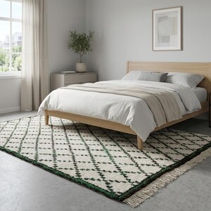 K&ouml;nnte beinhalten: Ein Schlafzimmer mit einem hellen Holzbettrahmen, wei&szlig;er Bettw&auml;sche und einem beigen &Uuml;berwurf. Auf dem Boden liegt ein gemusterter Teppich mit wei&szlig;er Basis und gr&uuml;nem Rautenmuster. Ein kleiner Nachttisch mit Lampe und gerahmtem Bild ist ebenfalls zu sehen.