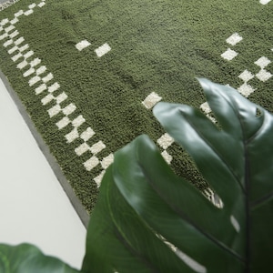 May include: Green shag rug with a white checkered border and scattered white squares. The rug has a textured surface and a fringe on one edge. The rug is a rectangular shape.