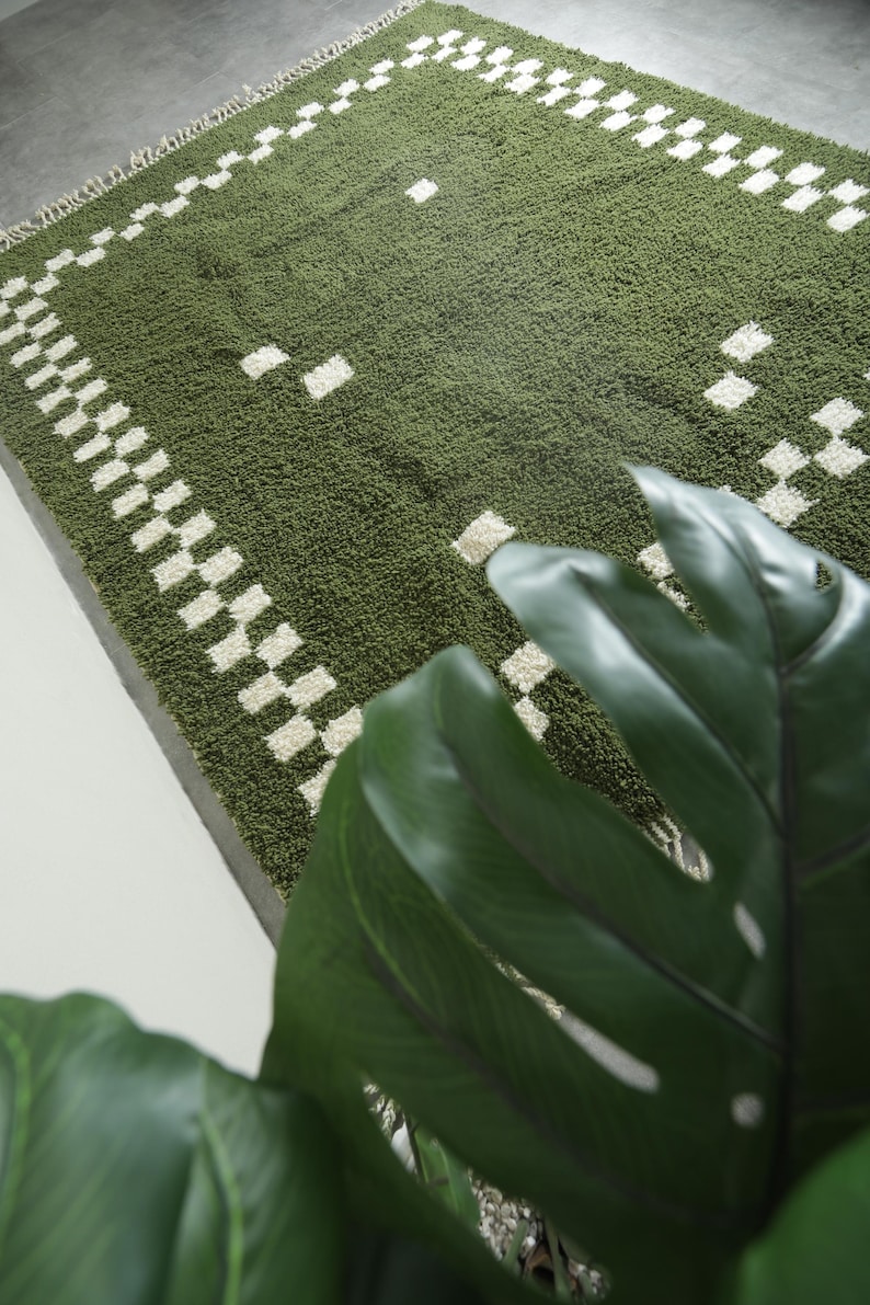 May include: Green shag rug with a white checkered border and scattered white squares. The rug has a textured surface and a fringe on one edge. The rug is a rectangular shape.