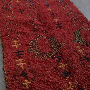 May include: A red Moroccan rug with a geometric pattern in black and gold. The rug has a thick pile and a fringe border.