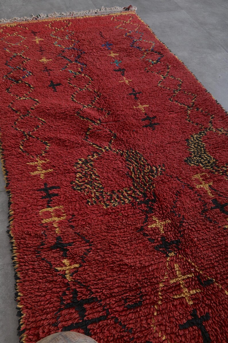 May include: A red Moroccan rug with a geometric pattern in black and gold. The rug has a thick pile and a fringe border.