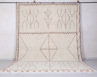 May include: A white Moroccan rug with a brown geometric pattern. The rug has a fringe border and is hanging on a wall.