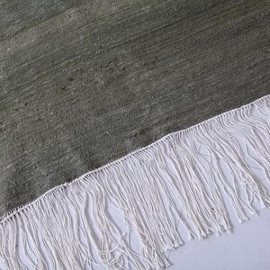 May include: A close-up of a green rug with a white fringe. The rug has a textured surface and a subtle sheen.