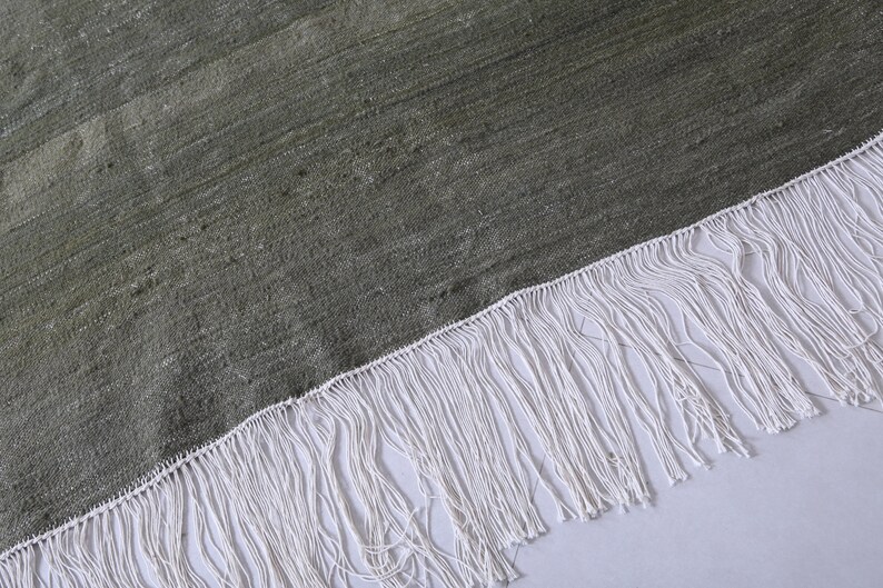 May include: A close-up of a green rug with a white fringe. The rug has a textured surface and a subtle sheen.