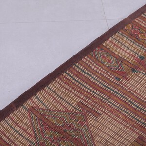 May include: A woven mat with a brown border. The mat features a repeating pattern of red, green, and yellow geometric shapes and stripes on a beige background. The mat is rectangular and appears to be made of natural fibers.