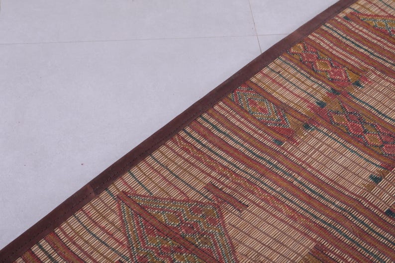 May include: A woven mat with a brown border. The mat features a repeating pattern of red, green, and yellow geometric shapes and stripes on a beige background. The mat is rectangular and appears to be made of natural fibers.