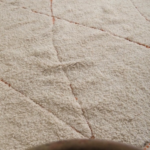 May include: A beige, textured rug with a geometric pattern. The rug features a diamond design with orange lines. The rug has a thick, plush pile, and the color is a warm, neutral tone, suitable for various interior styles.