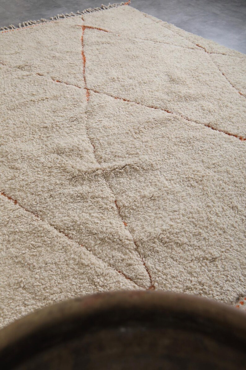May include: A beige, textured rug with a geometric pattern. The rug features a diamond design with orange lines. The rug has a thick, plush pile, and the color is a warm, neutral tone, suitable for various interior styles.