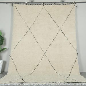 May include: Cream-coloured Moroccan rug featuring a black diamond pattern. The rug has a thick pile and a fringed edge. It is displayed against a neutral wall, with a plant in a white pot to the left.