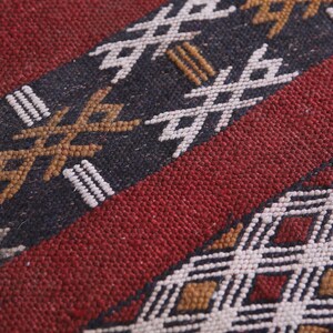 May include: Close-up of a red, black, and white woven rug with a geometric pattern. The rug has a textured surface and is made of natural fibers.