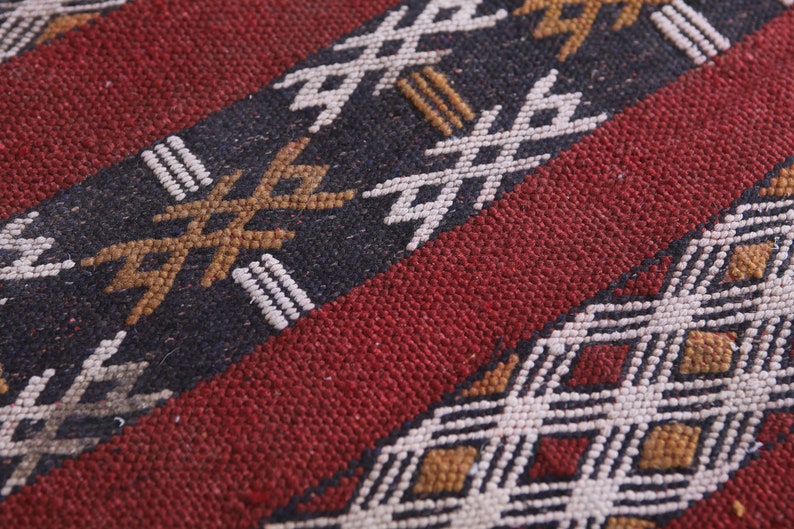 May include: Close-up of a red, black, and white woven rug with a geometric pattern. The rug has a textured surface and is made of natural fibers.