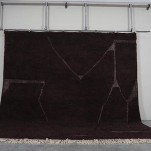 May include: A large, dark brown rug with a geometric design in lighter tones. The rug has a shaggy texture and a cream-colored fringe along the bottom edge. It is hanging from a metal bar.
