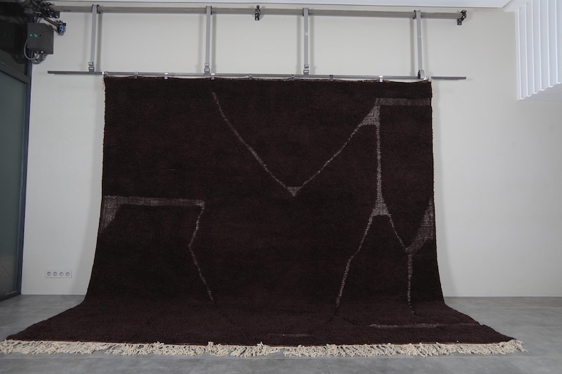 May include: A large, dark brown rug with a geometric design in lighter tones. The rug has a shaggy texture and a cream-colored fringe along the bottom edge. It is hanging from a metal bar.