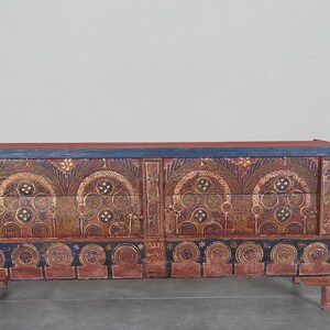 May include: An antique wooden chest with a red and blue painted finish. The chest features intricate, symmetrical gold and brown designs. The top and base have a blue border, and it rests on short, rounded legs. A decorative storage piece.