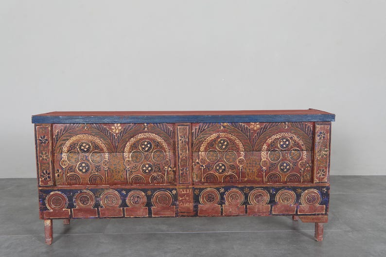 May include: An antique wooden chest with a red and blue painted finish. The chest features intricate, symmetrical gold and brown designs. The top and base have a blue border, and it rests on short, rounded legs. A decorative storage piece.