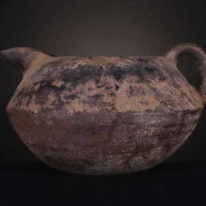 May include: A brown, round, ceramic pitcher with a handle and a spout. The pitcher has a rough, textured surface and appears to be very old.