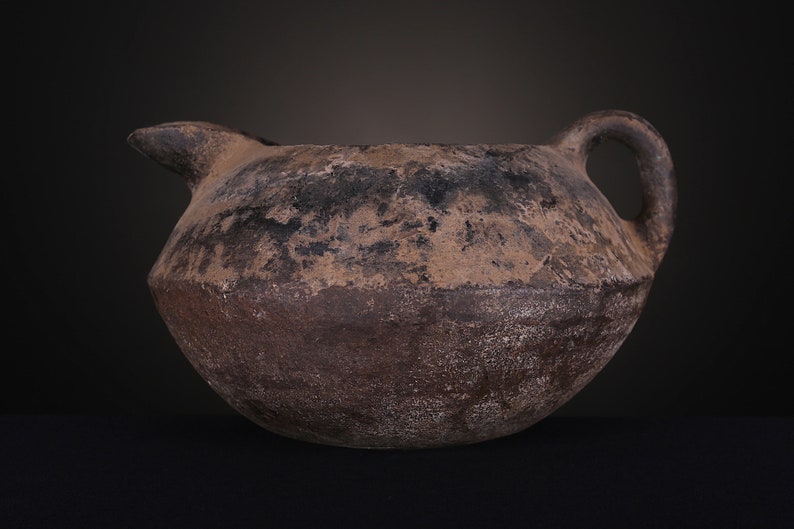 May include: A brown, round, ceramic pitcher with a handle and a spout. The pitcher has a rough, textured surface and appears to be very old.