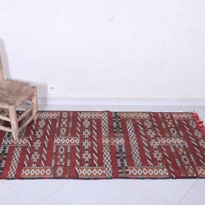 May include: A red, black, and white patterned rug with fringe on the ends. The rug is laid out on a white tiled floor with a wooden chair in the background.