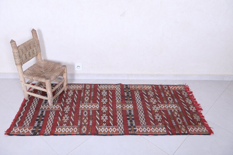 May include: A red, black, and white patterned rug with fringe on the ends. The rug is laid out on a white tiled floor with a wooden chair in the background.