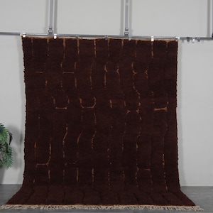 May include: A large, rectangular brown rug with a textured surface and a geometric pattern. The rug is displayed against a neutral wall, with a plant in a white pot to the left. The rug's dimensions are not visible.