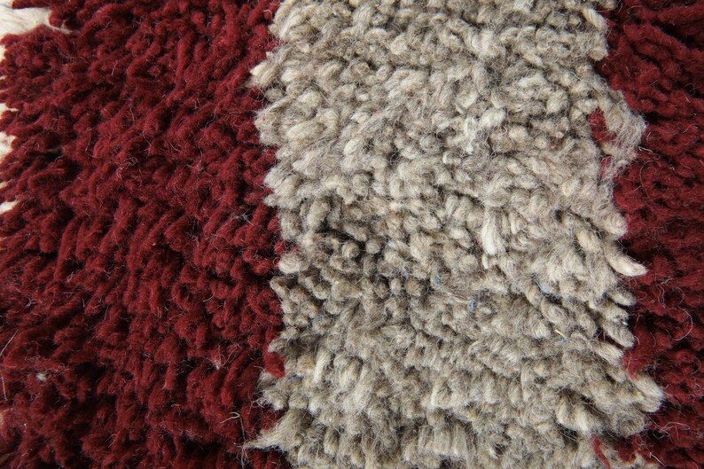 May include: Close-up of a textured rug with a thick, shaggy pile. The rug features a bold design with a deep burgundy red on the left and right sides, and a wide stripe of light gray in the center. The fibers are dense and create a soft, inviting look.
