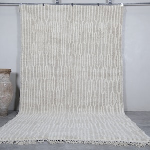 May include: A white and gray patterned rug with a fringe border. The rug is hanging on a wall and is made of a soft, fluffy material.