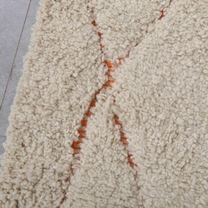 May include: Cream-colored, textured rug with a thick pile. The rug features a geometric design in orange, creating a striking contrast against the neutral background. The rug's plush texture suggests comfort and warmth, suitable for a living space.