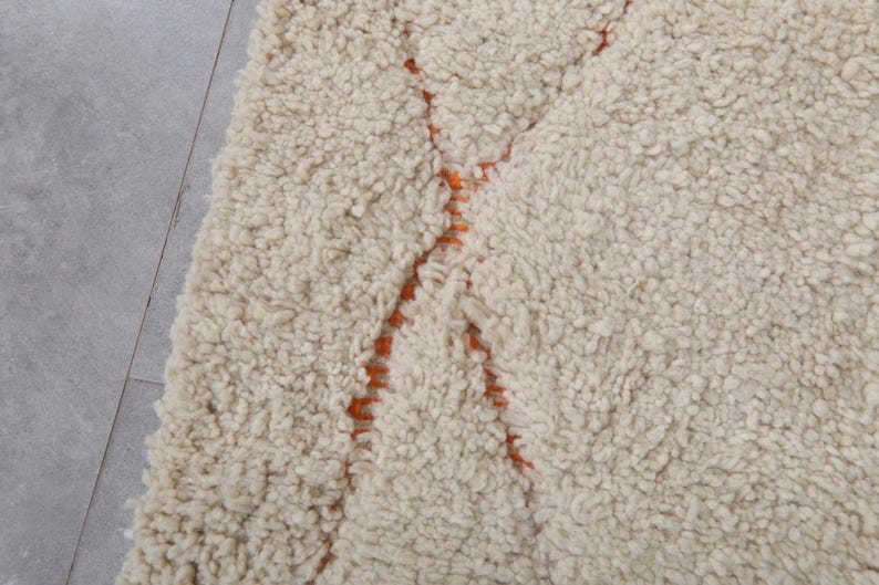 May include: Cream-colored, textured rug with a thick pile. The rug features a geometric design in orange, creating a striking contrast against the neutral background. The rug's plush texture suggests comfort and warmth, suitable for a living space.