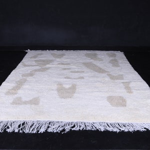 May include: A white rug with a beige geometric pattern. The rug has a fringe border.