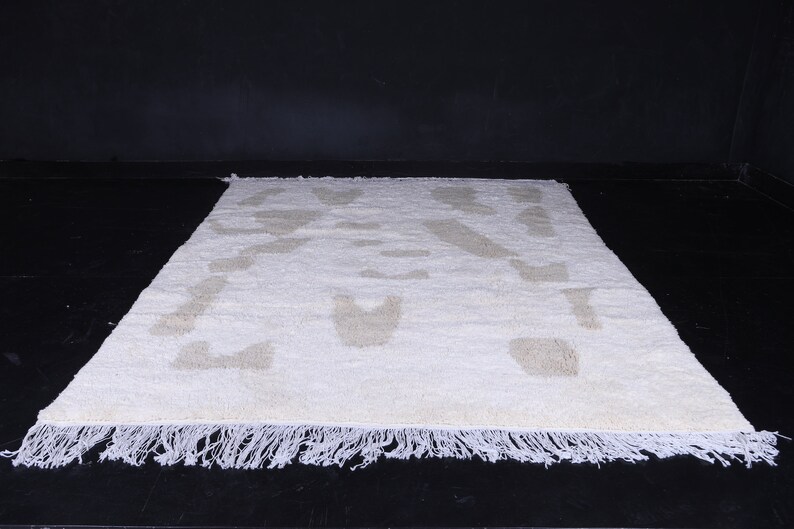May include: A white rug with a beige geometric pattern. The rug has a fringe border.