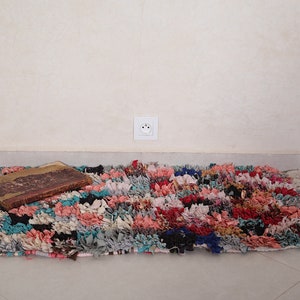 Puede incluir: Una alfombra colorida tejida a mano con materiales reciclados. La alfombra tiene una variedad de colores, incluyendo rosa, azul, verde y rojo. Est&aacute; colocada sobre un suelo de baldosas con un libro encima.