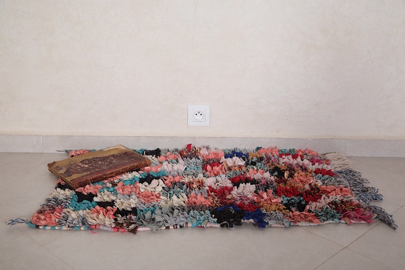 Puede incluir: Una alfombra colorida tejida a mano con materiales reciclados. La alfombra tiene una variedad de colores, incluyendo rosa, azul, verde y rojo. Est&aacute; colocada sobre un suelo de baldosas con un libro encima.