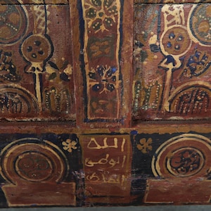 May include: Detailed view of a wooden panel with ornate, hand-painted patterns. The design incorporates circular and floral elements in brown, gold, and blue hues. Arabic script is present. The style indicates a historical or cultural object, possibly antique.