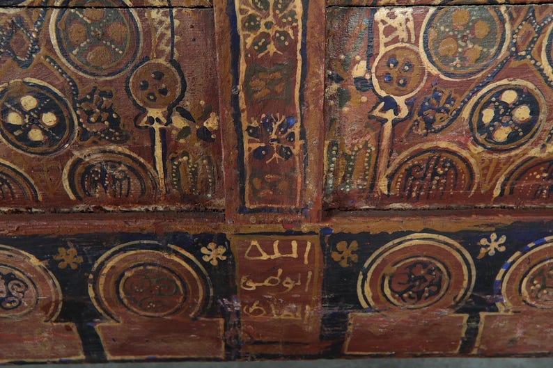 May include: Detailed view of a wooden panel with ornate, hand-painted patterns. The design incorporates circular and floral elements in brown, gold, and blue hues. Arabic script is present. The style indicates a historical or cultural object, possibly antique.