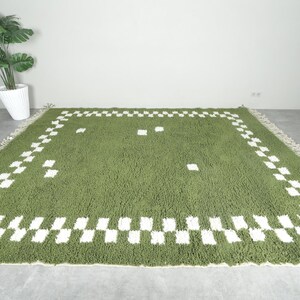 May include: A green rectangular rug with a white checkered border and scattered white squares. The rug has a shaggy texture and a fringe along the edges. The rug is placed on a gray concrete floor.