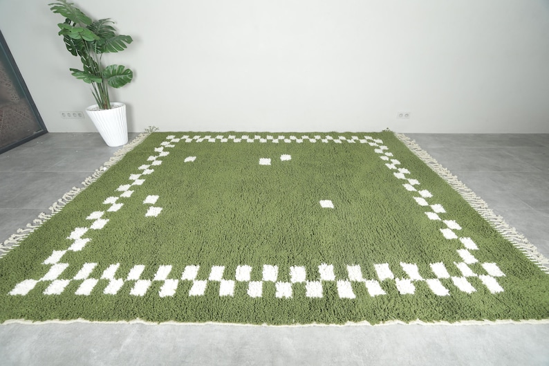 May include: A green rectangular rug with a white checkered border and scattered white squares. The rug has a shaggy texture and a fringe along the edges. The rug is placed on a gray concrete floor.