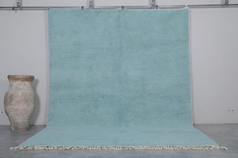 May include: A light blue shag rug with white fringe. The rug is hanging on a wall and is shown in a room with a white wall and a white vase.