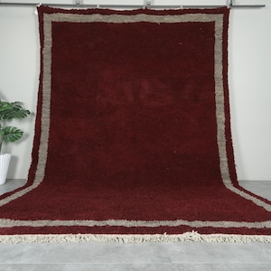 May include: A large, rectangular rug with a deep burgundy field and a wide, light gray border. The rug has a shaggy texture and a fringe along the edges. The rug is displayed against a white wall.