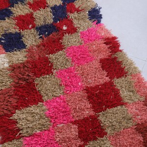 May include: A colourful, hand-woven rug with a geometric pattern in shades of red, pink, brown, and blue. The rug is made of thick, fluffy fibres and has a unique, textured look.