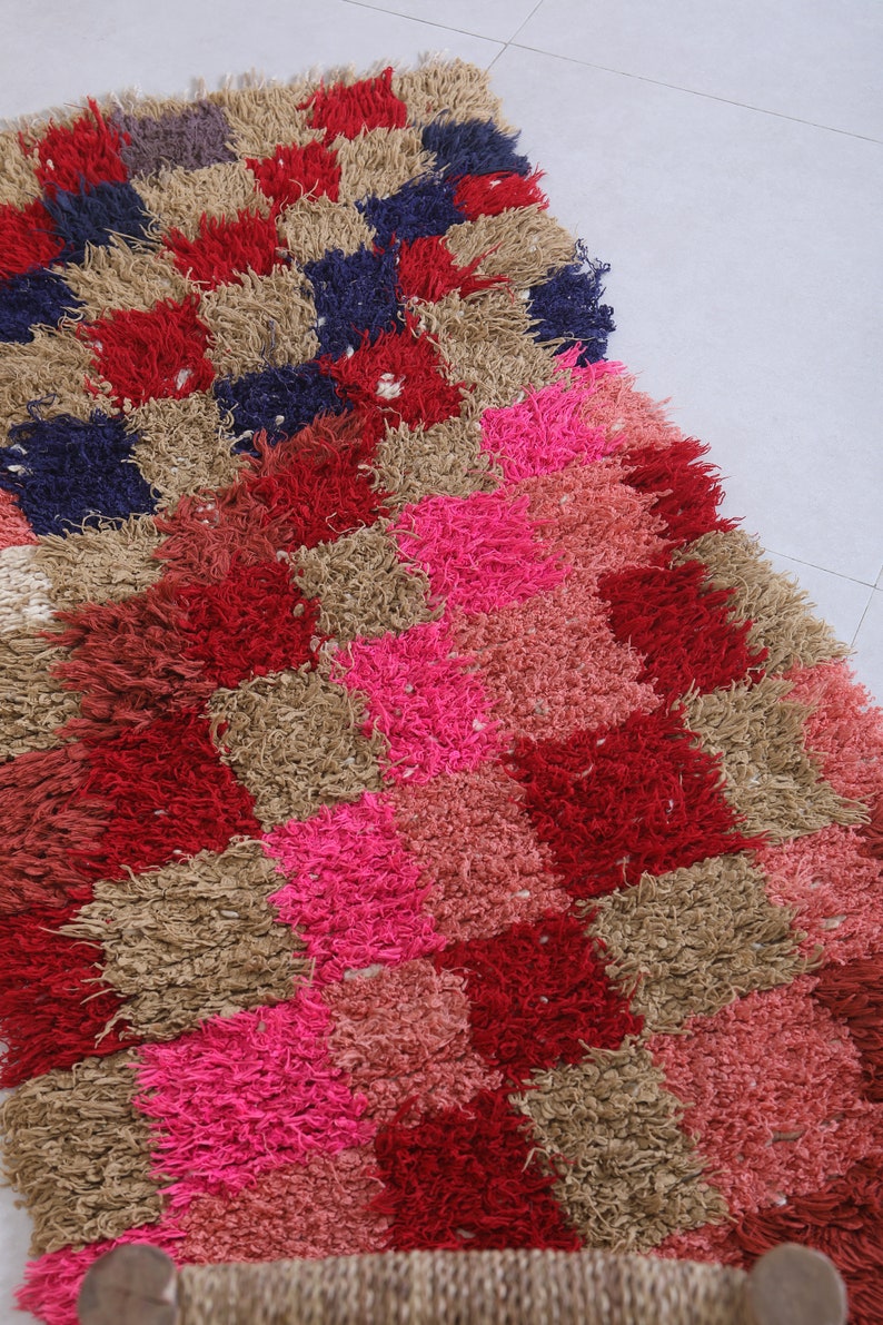 May include: A colourful, hand-woven rug with a geometric pattern in shades of red, pink, brown, and blue. The rug is made of thick, fluffy fibres and has a unique, textured look.