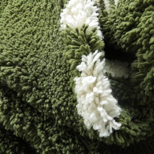 May include: Close-up of a green, textured rug with white, fluffy accents. The rug has a thick, shaggy pile, and the white accents are arranged in a striped pattern. The rug appears soft and inviting.