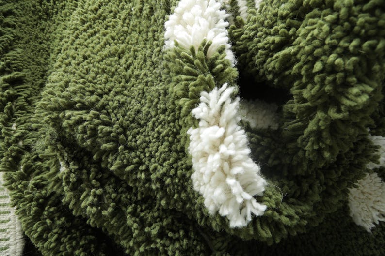 May include: Close-up of a green, textured rug with white, fluffy accents. The rug has a thick, shaggy pile, and the white accents are arranged in a striped pattern. The rug appears soft and inviting.