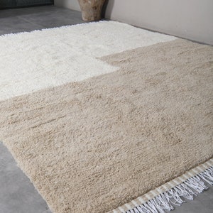 Puede incluir: Una alfombra beige y blanca con un borde de flecos. La alfombra est&aacute; hecha de un material suave y esponjoso.