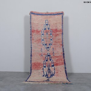 May include: A long, narrow rug with a pink and white geometric pattern. The rug has a blue border and is made of wool. It measures 3 feet by 7.5 feet.