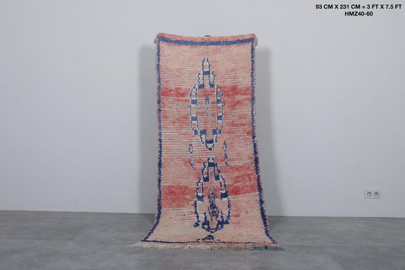 May include: A long, narrow rug with a pink and white geometric pattern. The rug has a blue border and is made of wool. It measures 3 feet by 7.5 feet.