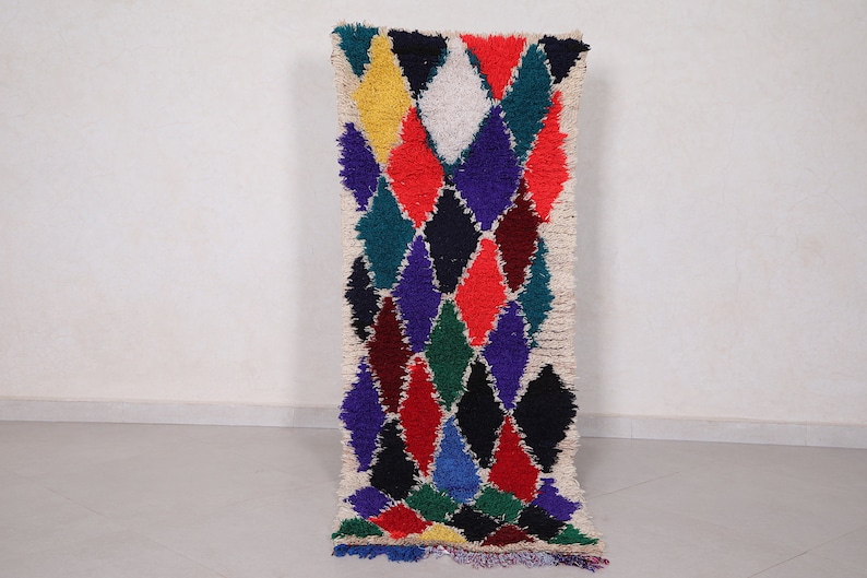 May include: A colorful Moroccan rug with a diamond pattern in shades of red, blue, green, yellow, and white. The rug has a fringe border and is made of wool.