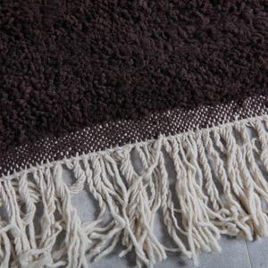 May include: Close-up of a brown, textured rug with a cream-colored fringe. The rug has a woven border in a lighter shade of brown. The fringe is made of thick, soft yarn.