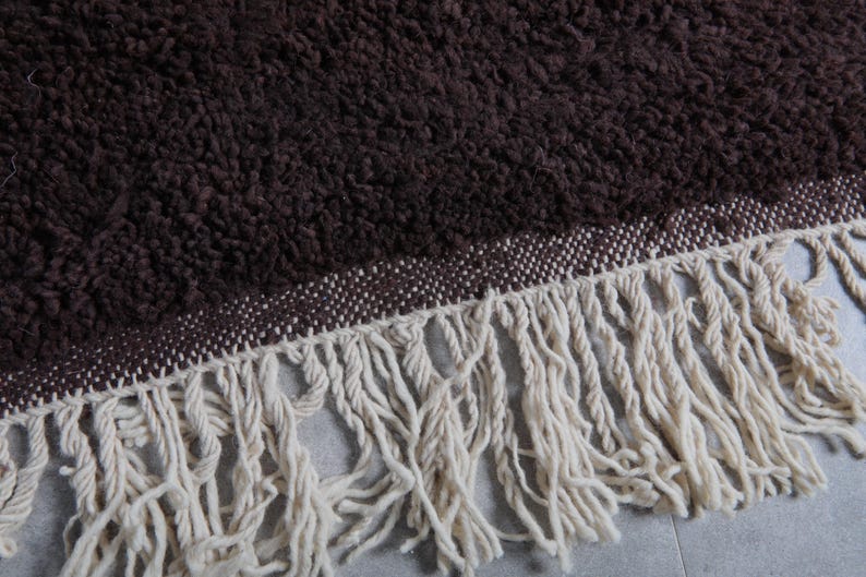 May include: Close-up of a brown, textured rug with a cream-colored fringe. The rug has a woven border in a lighter shade of brown. The fringe is made of thick, soft yarn.