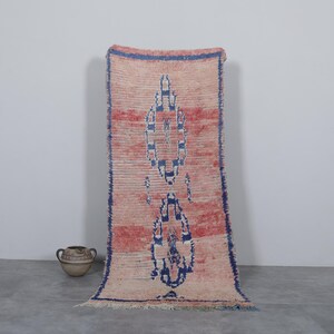May include: A long, rectangular rug with a faded pink and white background. The rug has a blue and white geometric pattern in the center.