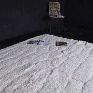 May include: A white fluffy rug with a textured pattern. The rug has a subtle wave-like design and is laid out on a dark floor.